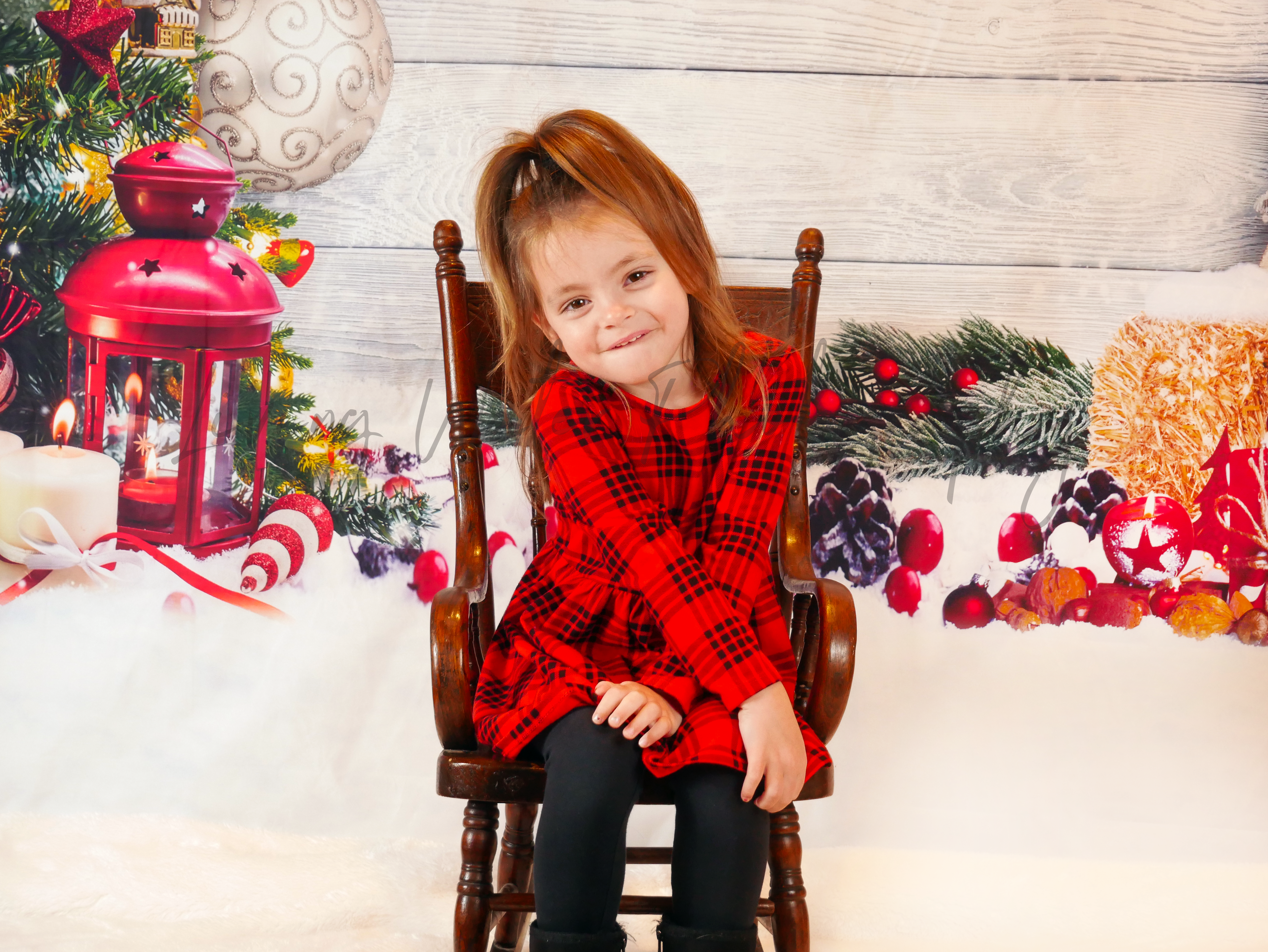 Young girl in red plaid dress seated in rocking chair with Christmas backdrop